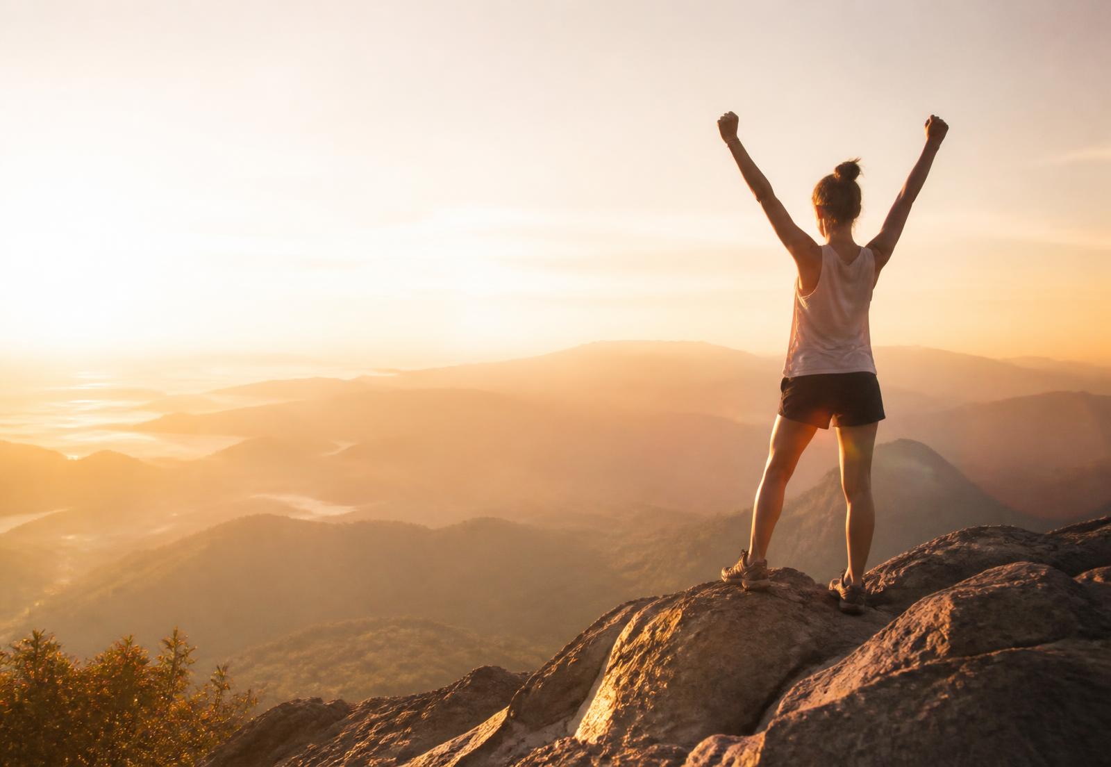 A woman on a mountain summit at sunrise, arms raised in triumph